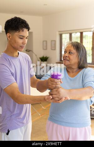 Il fisioterapista maschile birazziale aiuta la paziente anziana con esercizi al braccio a casa, utilizzando i manubri. Sono in un luminoso e moderno soggiorno con Foto Stock