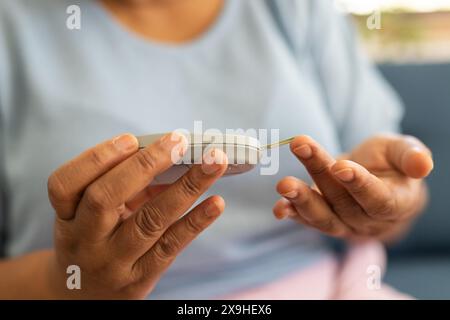 A casa, donna anziana birazziale che controlla zucchero nel sangue con glucometro. Seduto sul divano in un soggiorno ben illuminato con sfondo beige, concentrandosi su di lei Foto Stock