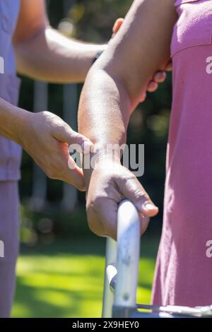 A casa, giovane infermiera maschile birazziale che aiuta paziente anziano con il braccio. Si trovano all'aperto in un giardino soleggiato e si concentrano sugli esercizi di riabilitazione Foto Stock