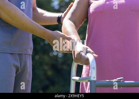 La giovane infermiera birazziale maschile aiuta la paziente anziana a deambulare a casa. Sono all'aperto in un giardino soleggiato, concentrandosi sull'esercizio di riabilitazione Foto Stock