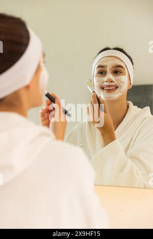 A casa, giovane donna caucasica che applica la maschera facciale mentre guarda allo specchio. Nel bagno accogliente, goditi una rilassante giornata in spa con illuminazione soffusa e fuliggine Foto Stock