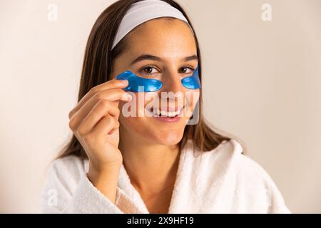 A casa, giovane donna caucasica che applica cerotti per gli occhi blu, sorridendo in accappatoio bianco. E' in un ambiente spa domestico luminoso e minimalista, godendo di un rel Foto Stock