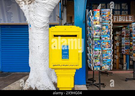 Essaouira, Marocco - 17 marzo 2024: Un negozio di souvenir colorato che vende cartoline accanto a una cassetta postale gialla brillante. Foto Stock