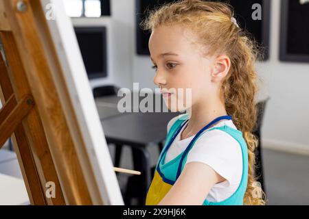 Ragazza caucasica con capelli biondi si concentra sulla pittura in una classe a scuola Foto Stock