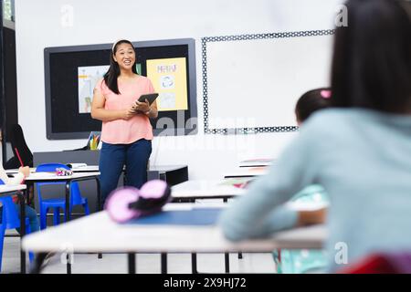 A scuola, giovane insegnante birazziale che tiene un tablet sorride in classe Foto Stock