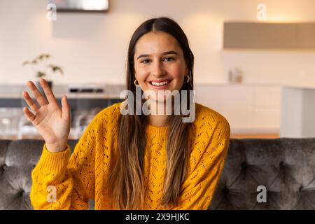 A casa, giovane donna caucasica con i capelli lunghi sorridente e saluta in videochiamata Foto Stock