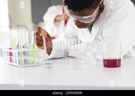 Un ragazzo birazziale adolescente conduce un esperimento in un laboratorio delle superiori. Indossato con un camice da laboratorio, analizza con attenzione una provetta, dimostrando una particolare attenzione alla S. Foto Stock