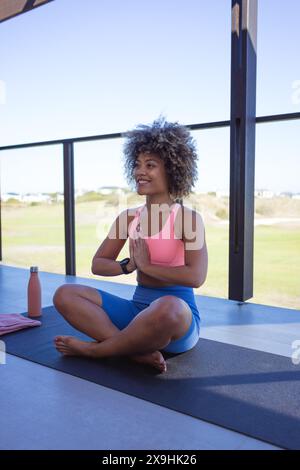 Sul balcone, una giovane donna birazziale che indossa il top rosa, leggings blu e pratica yoga. Seduti a gambe incrociate con capelli marroni ricci, godendosi la tranquillità del mese Foto Stock