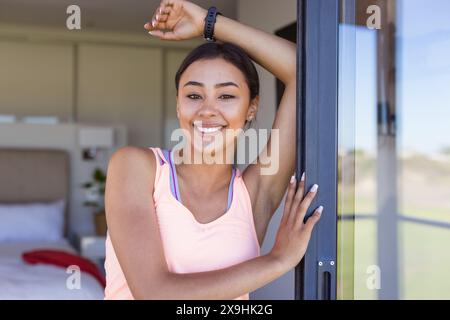 Una giovane donna birazziale a casa, che indossa la parte superiore rosa, che si appoggia su un telaio della porta, sorride Foto Stock