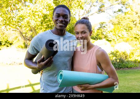 Una coppia giovane e variegata che tiene stuoie per lo yoga, sorridendo all'aperto Foto Stock