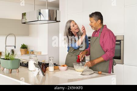 A casa, diverse coppie senior che cucinano insieme in una cucina moderna, indossando grembiuli. Donna caucasica con lunghi capelli grigi e rughe che toccano giocosamente Foto Stock