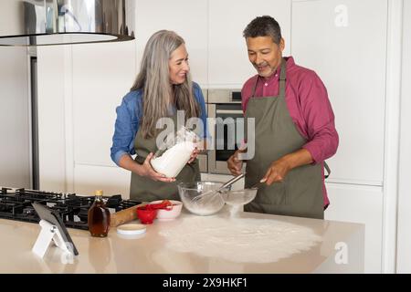 Diverse coppie di anziani sono felici di cucinare a casa in una cucina moderna. Donna caucasica con lunghi capelli grigi che reggono il vaso della farina, uomo birazziale con grigio corto Foto Stock