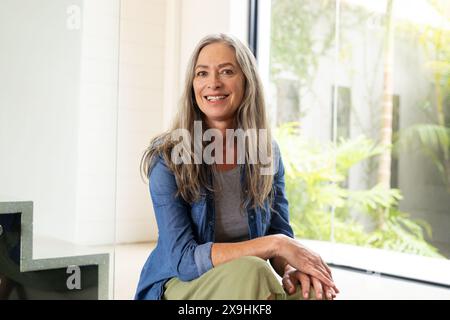 A casa, donna caucasica anziana con lunghi capelli grigi e occhi blu sorridenti all'interno. Indossando una camicia blu sopra la parte superiore grigia e pantaloni verdi, seduti vicino a vincere Foto Stock