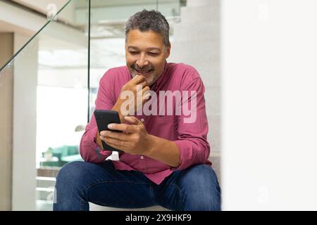 A casa, un maschio senior birazziale con capelli ricci corti e barba si siede sulle scale. Indossa una camicia rosa e jeans blu, sorridendo mentre usa il suo telefono, unal Foto Stock