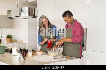 A casa, diverse coppie anziane indossano grembiuli da cuocere insieme in una cucina moderna. Donna caucasica con lunghi capelli grigi e uomo birazziale con breve buio Foto Stock
