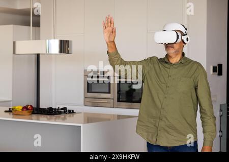 A casa, un uomo anziano birazziale indossa un visore per la realtà virtuale in una cucina moderna, spazio di copia. Coinvolgimento in un ambiente virtuale vicino a Kitchen Island Foto Stock