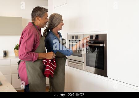 A casa, diverse coppie anziane usano il forno, sia indossando grembiuli che sorridendo. Marito birazziale di mezza età con capelli grigi e moglie caucasica con kit in mano Foto Stock