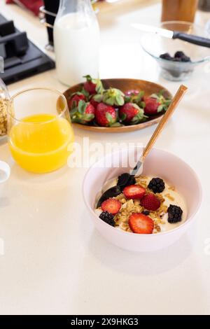 Colazione casalinga: Ciotola di muesli con yogurt, fragole e more, spazio fotocopie. Succo d'arancia in bicchiere, bottiglia di latte e piatto di fragole Foto Stock