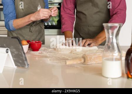 A casa, diverse coppie anziane in grembiuli che cucinano insieme in cucina. Sono circondati da utensili da forno, ingredienti e una tavoletta per seguire un re Foto Stock