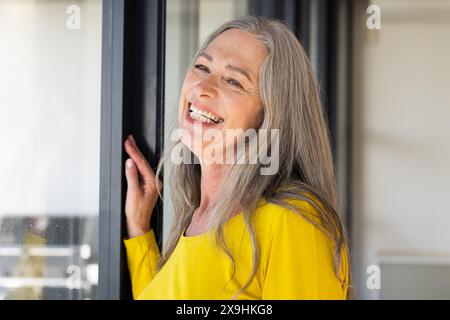 A casa, donna anziana caucasica con lunghi capelli grigi e rughe sorridenti alla finestra. Indossa un top giallo brillante, che sembra felice e rilassato in un buco Foto Stock