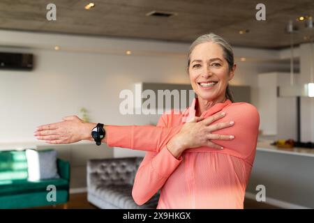 A casa, donna anziana caucasica con capelli grigi e occhi blu che si allungano. Indossando un top atletico rosa a manica lunga e un orologio fitness nero, sorridono Foto Stock