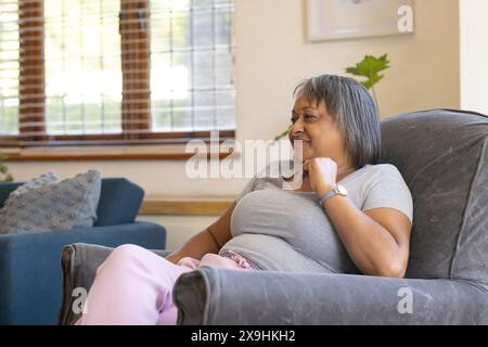 A casa, donna anziana birazziale seduta comodamente su una sedia, che sembra rilassata. La luce del sole filtra attraverso le tende da finestra, illuminando un accogliente salotto Foto Stock
