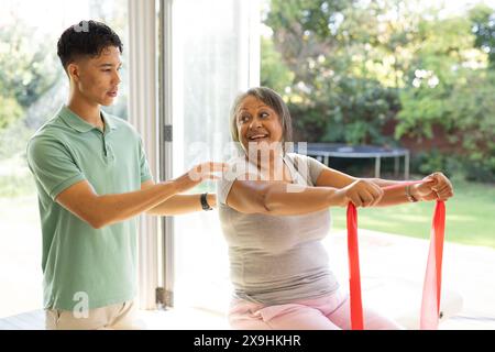A casa, fisioterapista maschile birazziale e paziente anziano che fa esercizi. Sono in camere luminose con grandi finestre che si affacciano su di un giardino verde Foto Stock