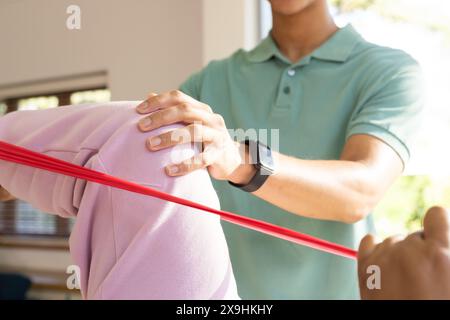 A casa, il giovane fisioterapista birazziale maschile assiste paziente anziano birazziale femminile. Clinica luminosa con grandi finestre e attrezzature moderne, utilizzando i servizi Foto Stock