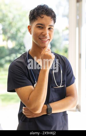 Infermiera maschile birazziale in scrub della marina con stetoscopio, sorridendo con sicurezza all'aperto. Clinica luminosa e ariosa con grandi finestre e vegetazione esterna, non alta Foto Stock