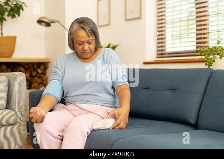 A casa, donna anziana birazziale che controlla la pressione sanguigna sul divano in soggiorno, copia spazio. Arredi moderni e luce naturale che riempie lo spazio per creare cal Foto Stock