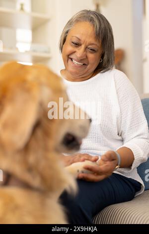 A casa, donna anziana birazziale sorride mentre interagisce con il cane su un divano Foto Stock