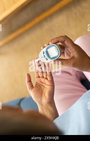 A casa, donna anziana birazziale che controlla i livelli di zucchero nel sangue con glucometro. Seduto sul divano in un accogliente soggiorno con pavimento in moquette e legno di fu Foto Stock