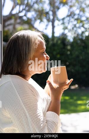 Donna birazziale anziana all'aperto che sorseggia una bevanda calda, avvolta in un comodo maglione bianco. La luce del sole filtra tra gli alberi in un giardino tranquillo, creando un ser Foto Stock