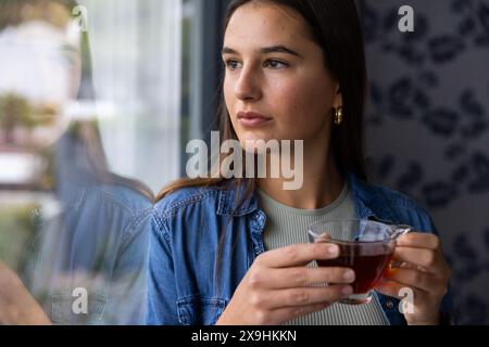 A casa, una giovane donna caucasica che tiene una tazza di tè, guarda con attenzione fuori dalla finestra. Si trova in un accogliente ambiente termale domestico con illuminazione soffusa e calma Foto Stock