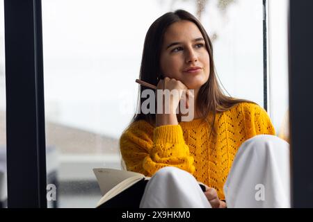 A casa, giovane donna caucasica che si rilassa con diario, indossando un comodo maglione giallo. Seduti vicino a una grande finestra, godendosi un momento di pace con un sereno Foto Stock