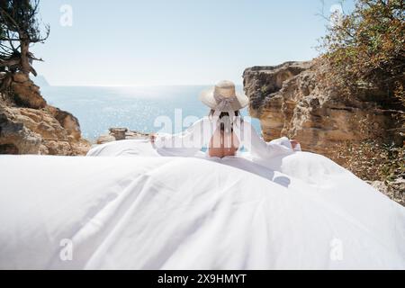 Una donna in abito bianco si trova su una scogliera rocciosa che si affaccia sull'oceano. Sta indossando un cappello di paglia e si sta godendo la vista. Foto Stock