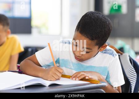 Ragazzo birazziale si è concentrato sulla scrittura in un taccuino con una matita in una classe scolastica Foto Stock