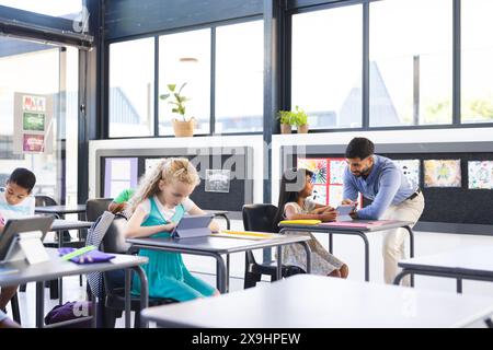 A scuola, diversi gruppi di studenti che imparano con il loro insegnante asiatico maschile in classe. Indossa una camicia blu, aiutando una donna birazziale Foto Stock