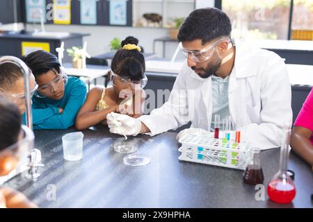 Il giovane insegnante asiatico conduce un esperimento scientifico con diversi bambini a scuola. Ragazze e ragazzi birazziali guardano con attenzione come dimostra in una Foto Stock