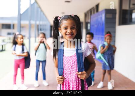 A scuola, la giovane ragazza birazziale con vestito rosa e zaino sorride all'aperto Foto Stock