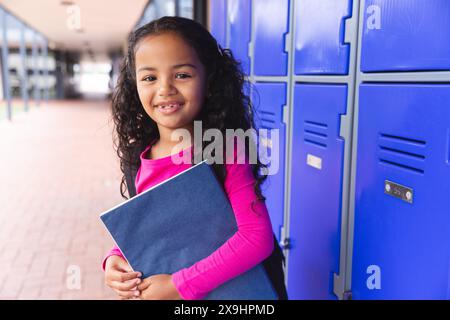 A scuola, all'aperto, una giovane ragazza birazziale con una cartella blu si trova accanto agli armadietti Foto Stock