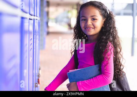 A scuola, giovane studentessa birazziale che tiene libri, in piedi accanto a armadietti all'aperto Foto Stock