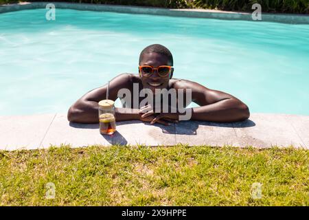 Un giovane afroamericano che si rilassa all'aperto in piscina, sorseggiando un drink Foto Stock