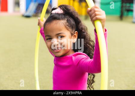 Nel parco giochi della scuola all'aperto, giovane ragazza birazziale che tiene i cerchi gialli e sorride Foto Stock