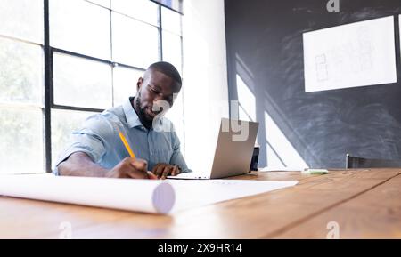 Al moderno ufficio commerciale, giovane afroamericano che lavora su disegni architettonici. Utilizzare una matita per disegnare su carta di grandi dimensioni mentre si guarda il notebook Foto Stock