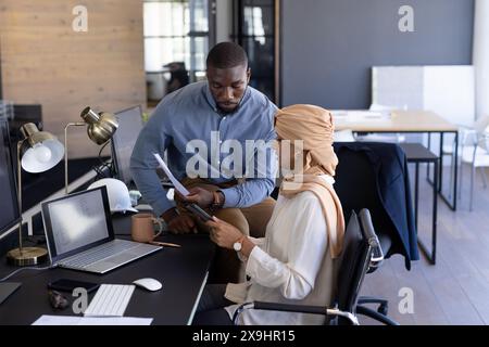 Nel moderno ufficio aziendale, un team diversificato che parla di documenti e utilizza un tablet. Uomo afroamericano in camicia blu e donna indiana in beige hijab col Foto Stock