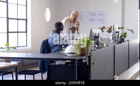 Team diversificato nel moderno ufficio aziendale che celebra il successo con il pugno. Uomo afroamericano in camicia blu seduto alla scrivania, leader di squadra asiatica Foto Stock
