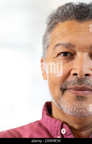 A casa, un maschio senior birazziale con capelli grigi corti e barba in piedi all'interno, spazio copia. Indossa una camicia rossa e guarda direttamente la fotocamera con un neu Foto Stock
