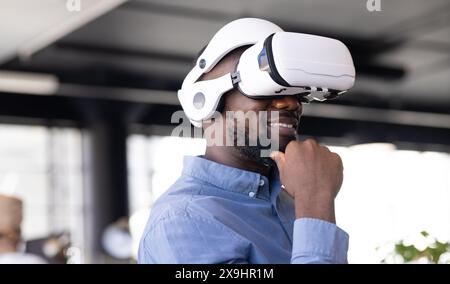Giovane afroamericano che indossa un visore per la realtà virtuale in ufficio. Ha i capelli neri corti, la barba e sorride mentre tocca il mento Foto Stock