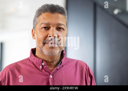 A casa, un maschio senior birazziale con capelli corti e ricci grigi e barba in piedi all'interno. Indossa una camicia rosa con bottoni e guarda la fotocamera con una calma Foto Stock